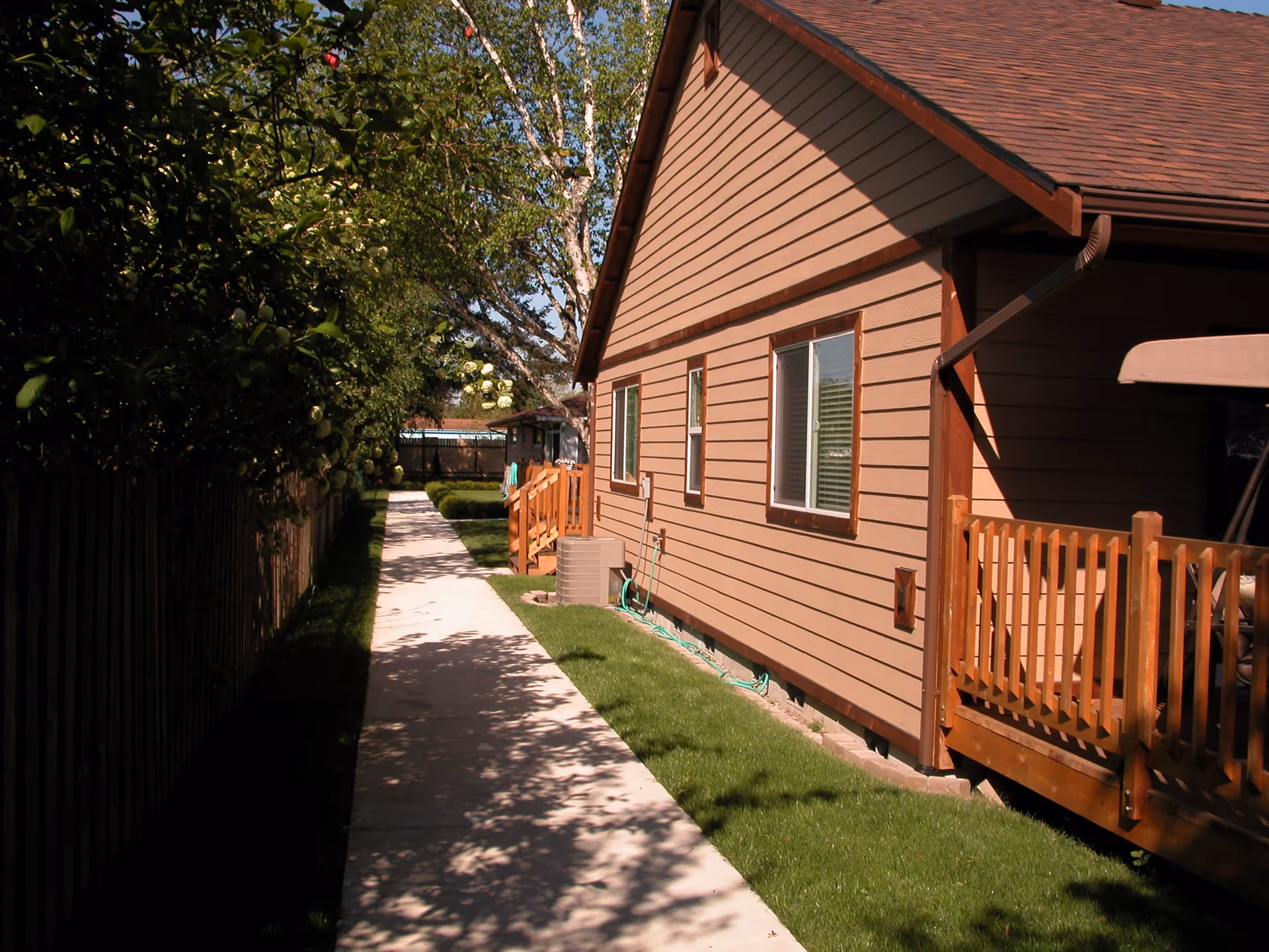 A narrow concrete pathway runs alongside a brown wooden building with a small wooden deck and railing. The building has two windows and a sloped roof. On the left side of the pathway, there is a wooden fence with green bushes and trees providing shade.