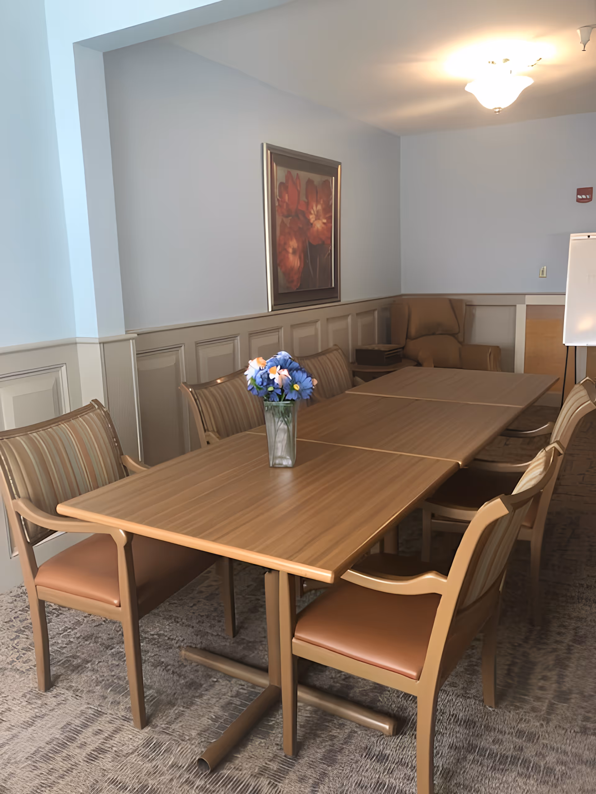 A bright dining/activity room with a long wooden table surrounded by chairs, a vase of blue flowers, wall art, and an armchair in the corner.