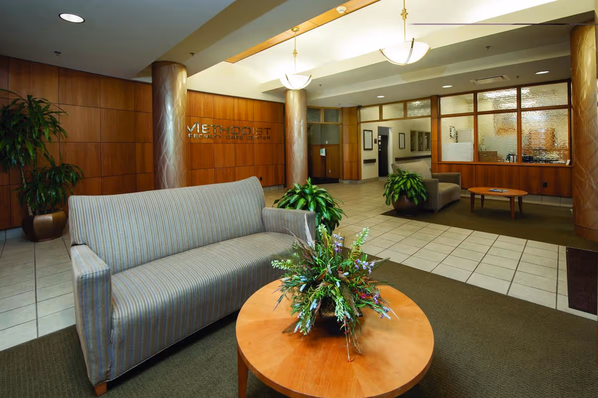 A spacious and well-lit lobby area of Methodist Specialty Care Center featuring a striped sofa, a round wooden coffee table with a floral arrangement, potted plants, wood-paneled walls, two large decorative columns, and a reception area with frosted glass windows in the background.