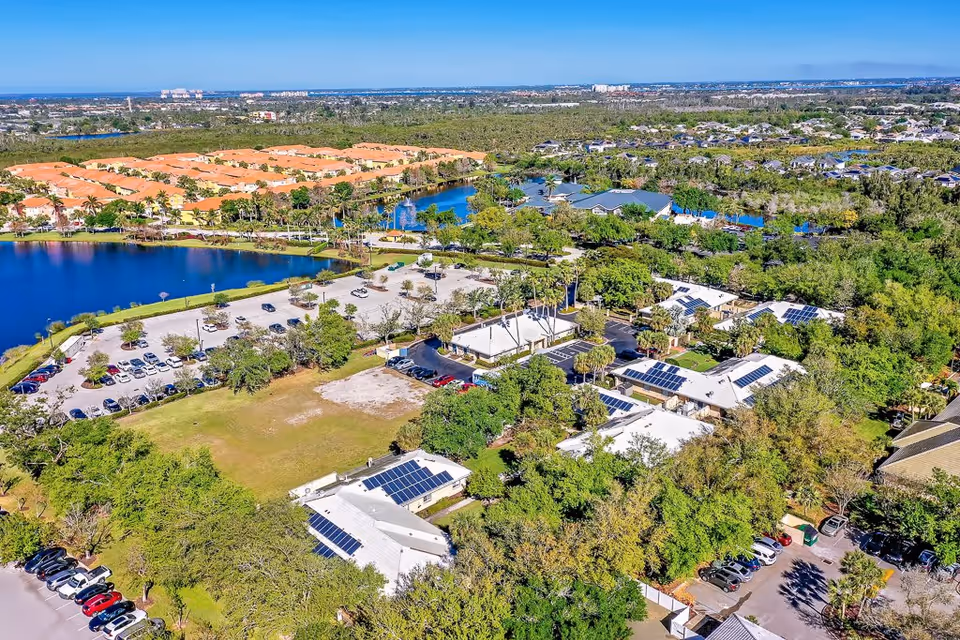 Aerial view of Healthpark Senior Living facility showing multiple buildings with white roofs and solar panels, surrounded by trees and greenery. There is a large parking lot with several cars parked, a blue lake with a fountain, and residential houses with orange roofs in the background under a clear blue sky.