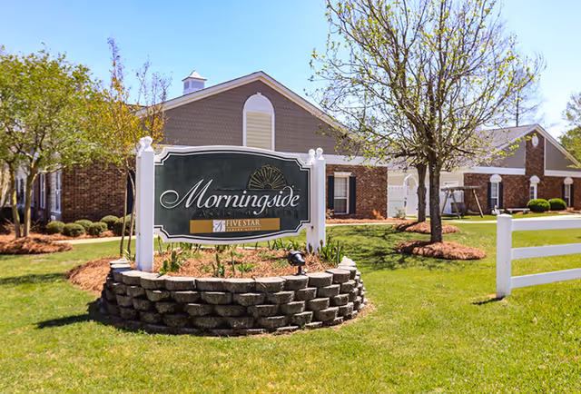 Exterior view of a senior living facility with a sign reading 'Morningside Assisted Living' surrounded by a circular stone planter, green grass, trees, and a white fence in front of a brick building under a clear blue sky.