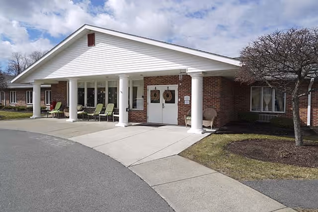 Exterior front view of a single-story brick building with white columns supporting a peaked roof over the entrance. There are chairs arranged on the porch area and a tree without leaves on the right side. The sky is partly cloudy.