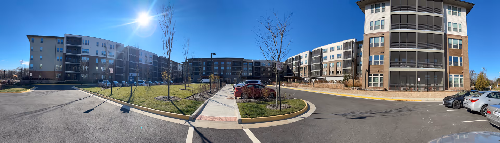 Panoramic exterior view of the Woodleigh Chase Senior Living complex showing multi-story buildings, a central walkway, parked cars, and a clear blue sky.