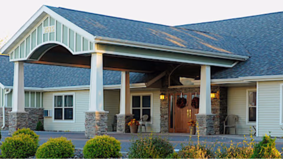 Covered front entrance of a senior living building with a porte-cochere, double wooden doors, stone columns, and shrubs.