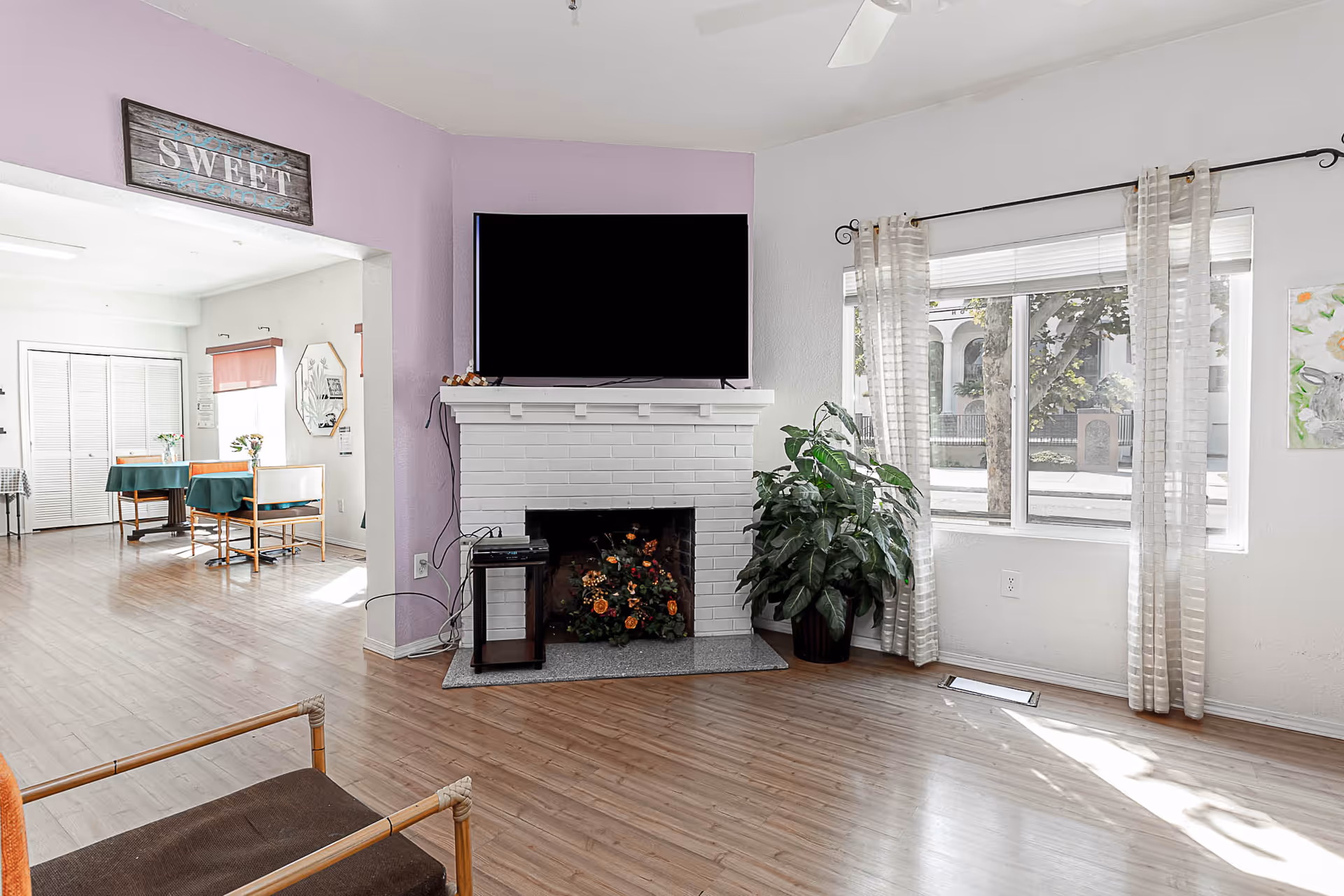 A bright living room with light wood flooring, a white brick fireplace with a flat-screen TV mounted above it, a large green plant next to the fireplace, and a window with sheer curtains letting in natural light. To the left, there is an open doorway leading to a dining area with tables and chairs, and a wall sign above the doorway that reads 'SWEET'.