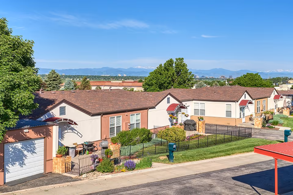 Row of single-story residential units with small fenced patios, landscaping, and distant mountain views under a clear blue sky.