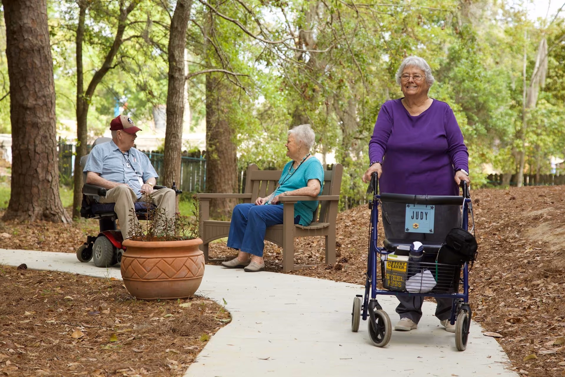 An elderly woman wearing a purple top uses a walker with a basket labeled 'Judy' while walking on a paved path in a wooded outdoor area. In the background, an elderly man in a motorized wheelchair and an elderly woman sitting on a bench are engaged in conversation. The scene is surrounded by trees and natural landscaping.