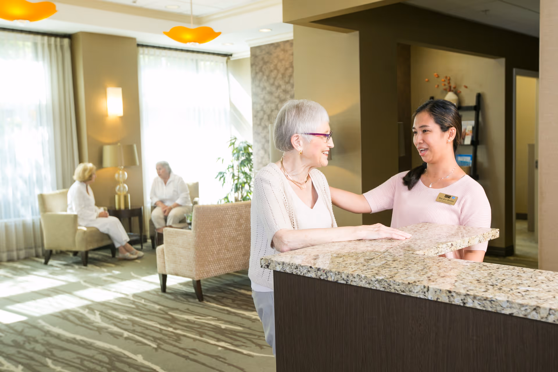 An elderly woman with white hair and glasses is smiling and talking to a female staff member wearing a pink shirt and name tag at a reception desk with a granite countertop in a well-lit common area. In the background, two elderly women are seated and conversing near large windows with sheer curtains, and the room is decorated with modern furniture and warm lighting.