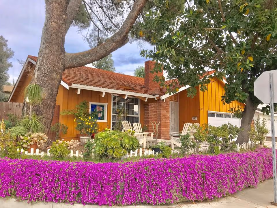 Exterior view of a single-story house with orange siding and a red tiled roof, surrounded by lush greenery and vibrant purple flowers along the sidewalk. There are white chairs on the front porch and large trees providing shade.