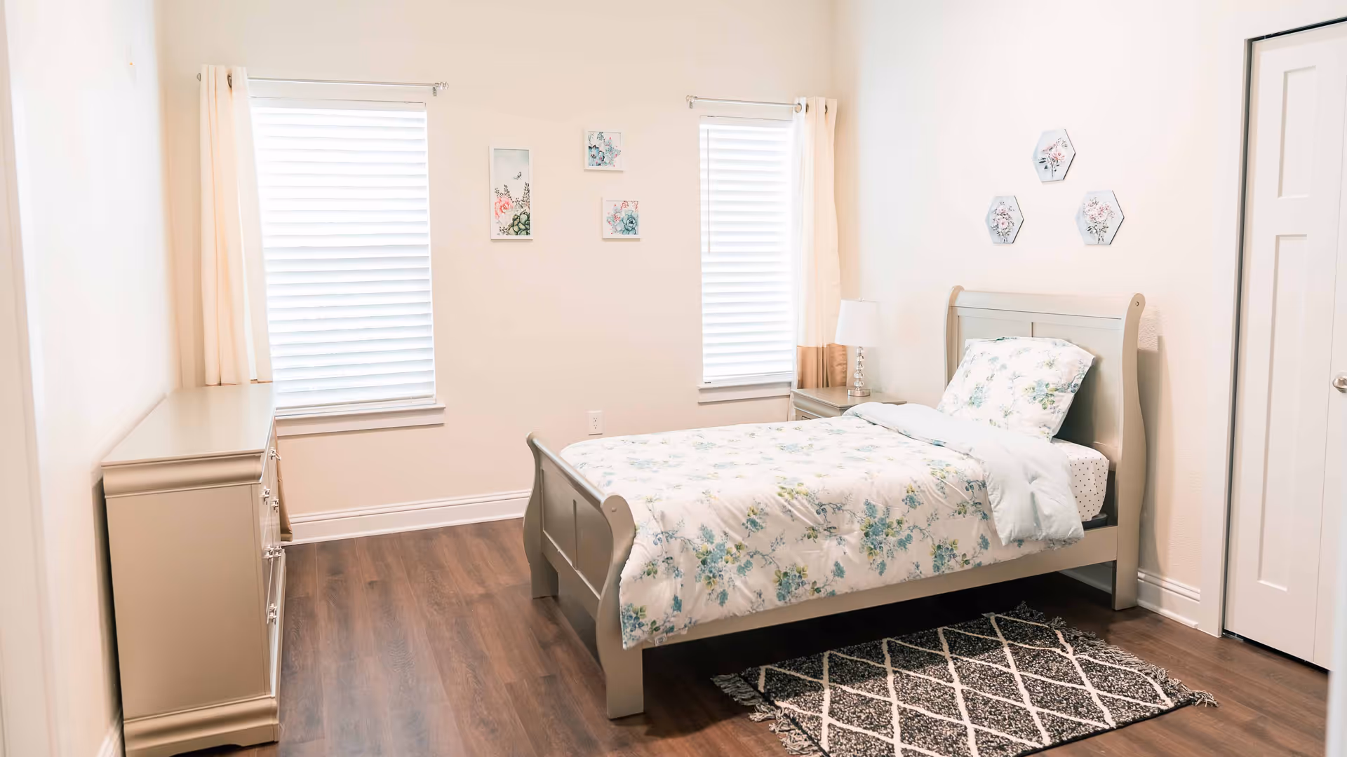 A bright and cozy bedroom with a single bed featuring floral bedding. The room has two windows with white blinds and light curtains, a nightstand with a lamp, a dresser, and floral wall decorations. The floor is wooden with a patterned rug beside the bed.