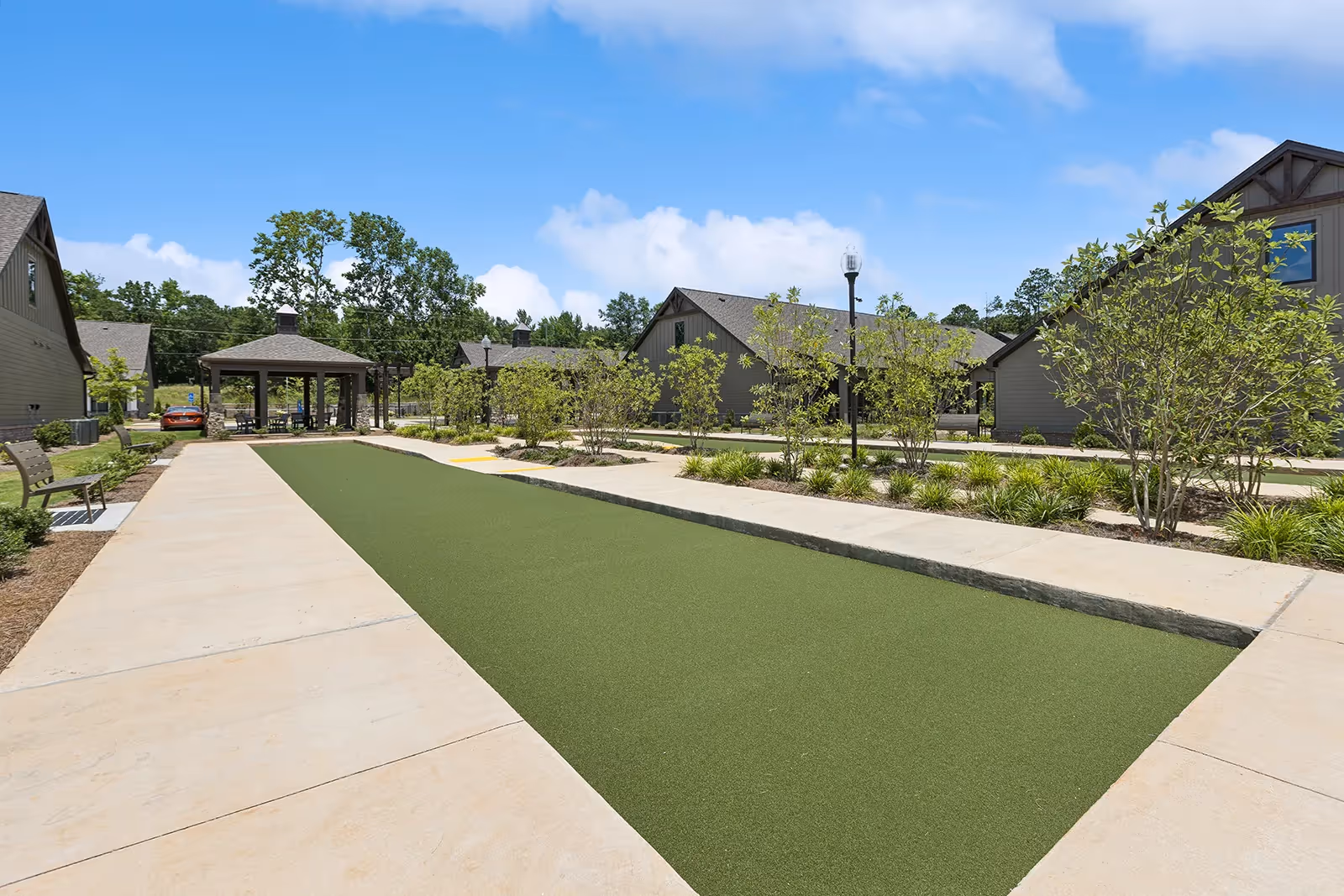 Outdoor area at The Phoenix at Opelika featuring a bocce ball court surrounded by concrete walkways, benches, small trees, and shrubs. Several buildings with gray siding and pitched roofs are visible in the background under a blue sky with some clouds.