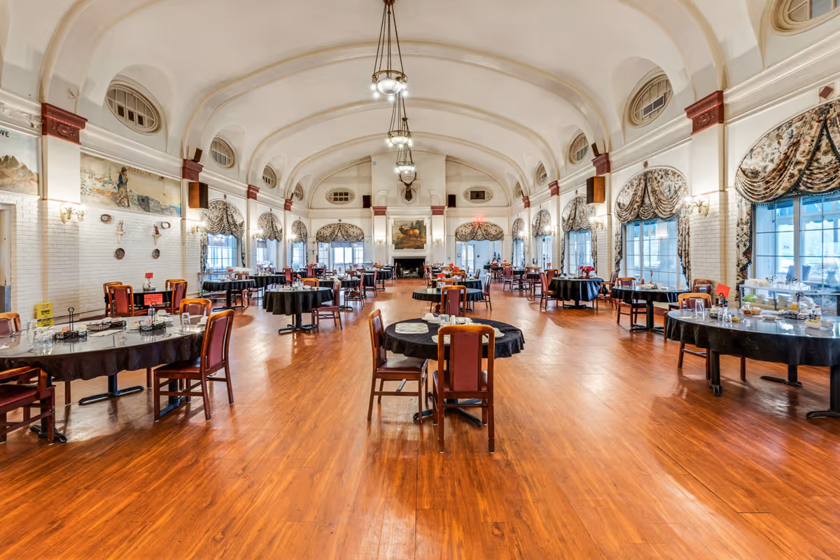 A spacious dining room with wooden floors and multiple round tables covered with black tablecloths. Each table is surrounded by wooden chairs with red cushions. The room features high arched ceilings with hanging chandeliers, large windows with decorative curtains, and a fireplace at the far end. The walls are white with red accents and decorated with paintings and wall sconces.