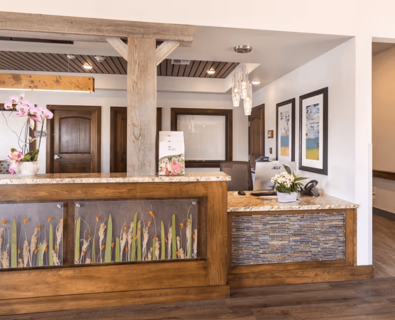 Reception desk area with wooden accents and decorative panels featuring grass designs. The desk has a granite countertop with a small potted plant and a computer. Behind the desk are two closed wooden doors and framed artwork on the wall. The floor is wooden and the ceiling has recessed lighting.