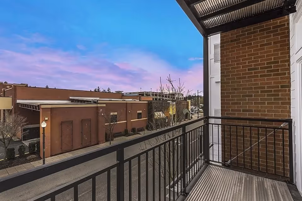 View from a balcony with black metal railing overlooking a street and a large brick building under a blue and purple sky at dusk.