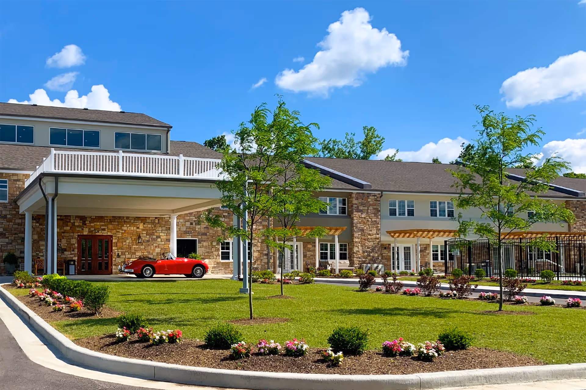 Exterior view of Sycamore Reserve Senior Living facility with a well-maintained lawn, flower beds, young trees, and a covered entrance. A red convertible car is parked under the entrance canopy. The building features stone and beige siding with multiple windows and a clear blue sky with some clouds in the background.