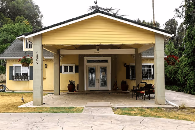 Front exterior view of a yellow single-story building with a covered entrance supported by two columns. The entrance has double glass doors with decorative glass panels. There are potted plants on either side of the doors and outdoor seating with chairs and a small table to the right. The building number 5809 is visible on the left column and above the entrance. Surrounding the building are trees and a lawn area.