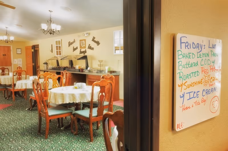 Dining room with round table and wooden chairs, a buffet station in the back, and a handwritten menu board on the right wall.