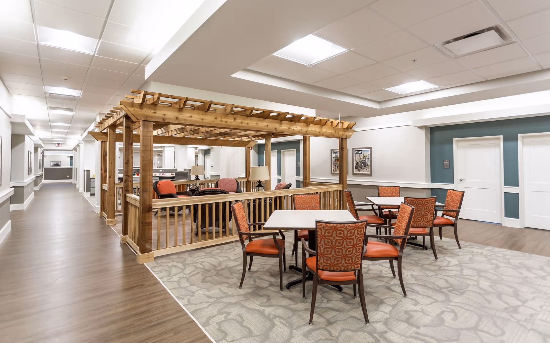 Interior view of a senior living facility common area at Heritage Crossing featuring a wooden pergola structure with cushioned chairs and tables arranged on a patterned carpet. The space has wood flooring, white walls with teal accent panels, framed artwork, and bright ceiling lights.