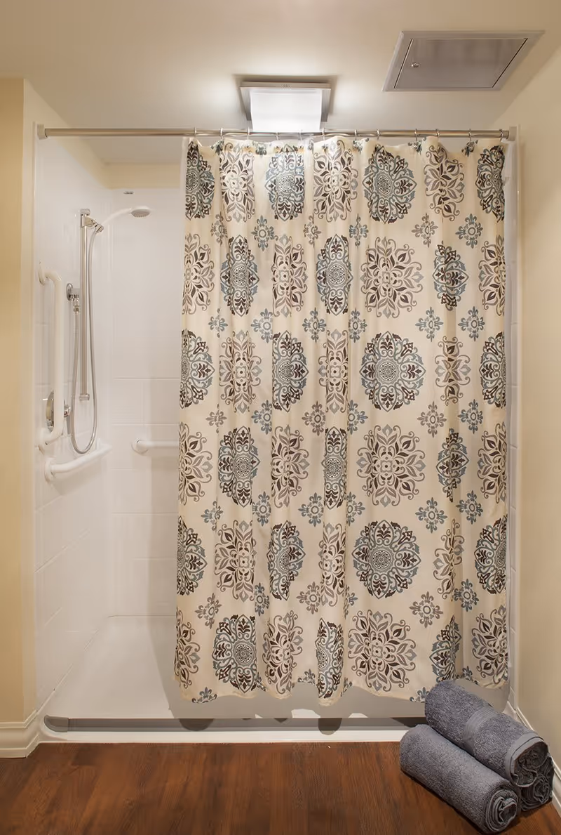 A shower area with a decorative patterned shower curtain featuring blue and beige designs. The shower has white tiled walls, a handheld showerhead, and grab bars for safety. Three rolled gray towels are stacked on the wooden floor outside the shower.