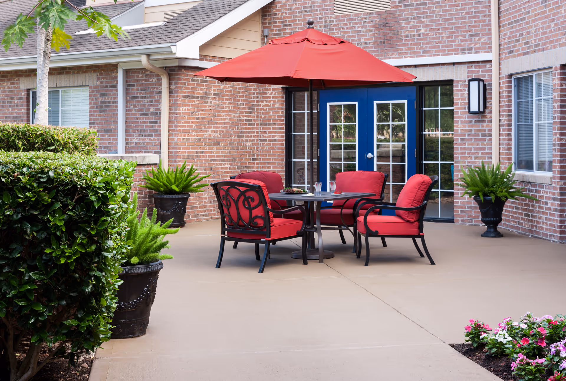 Outdoor patio area with a round table and four red cushioned chairs under a large red umbrella. The patio is surrounded by brick walls with blue double doors and windows. There are several potted green plants and trimmed bushes around the patio.
