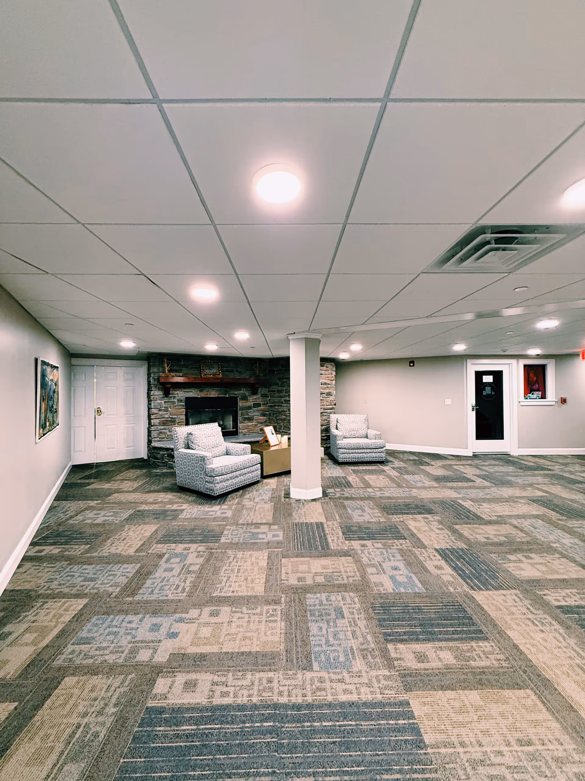 A spacious room with patterned carpet flooring, two cushioned armchairs facing a stone fireplace with a wooden mantle, a small table between the chairs, and a white column in the center. The walls are light-colored with a painting on the left wall, and there are multiple ceiling lights illuminating the room. A door and a window are visible on the right side.