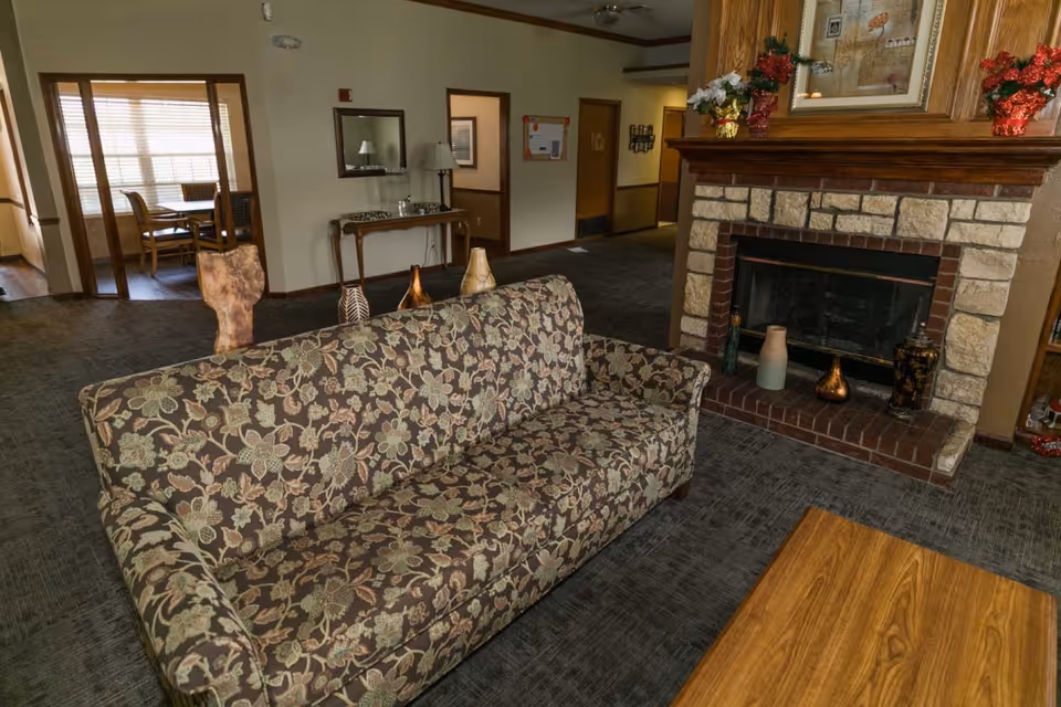 A cozy living room area in Meadow Creek Senior Living featuring a floral patterned sofa, a stone and brick fireplace with decorative vases on the hearth and mantel, and a wooden coffee table. In the background, there is a hallway with doors and a small table with a mirror above it, as well as a dining area with a table and chairs near a window with blinds.