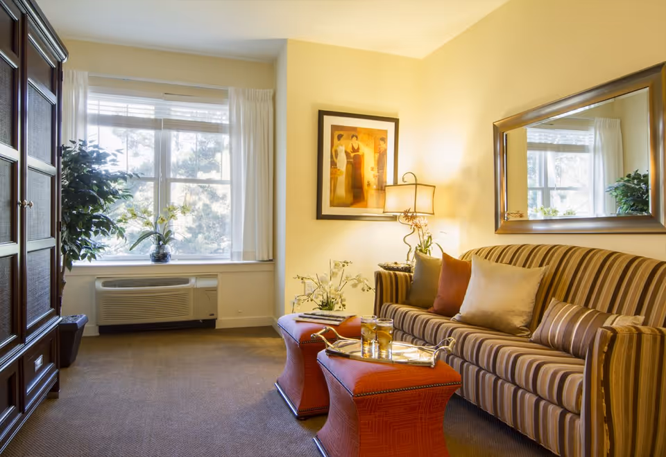 Sunlit sitting area with a striped sofa, red ottomans, a framed mirror, lamp and a window with potted plants.