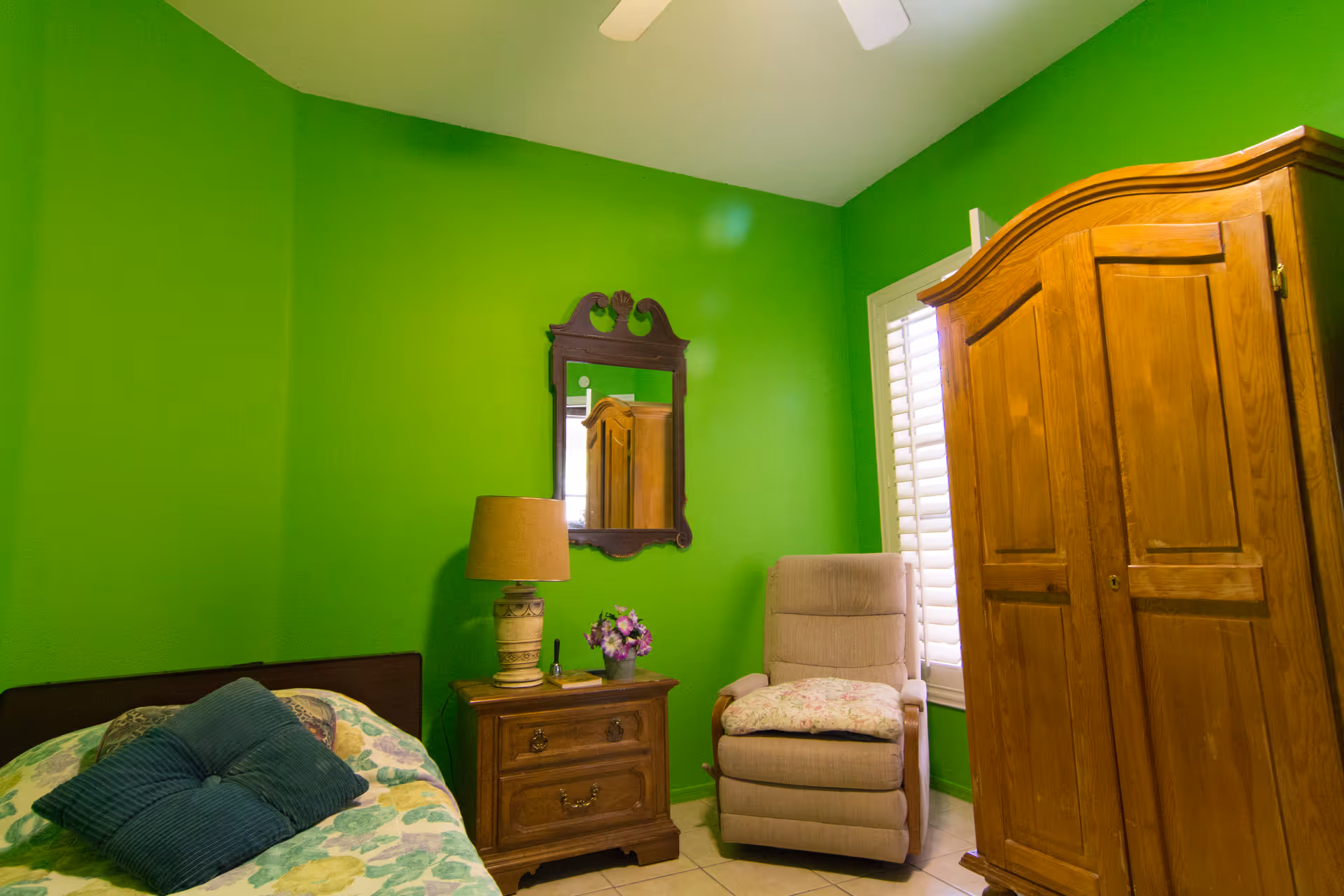 A bedroom with bright green walls featuring a bed with floral bedding and blue pillows, a wooden nightstand with a lamp and a small flower pot, a beige cushioned armchair with a floral cushion, a wooden wardrobe, a decorative wall mirror, and a window with white shutters.