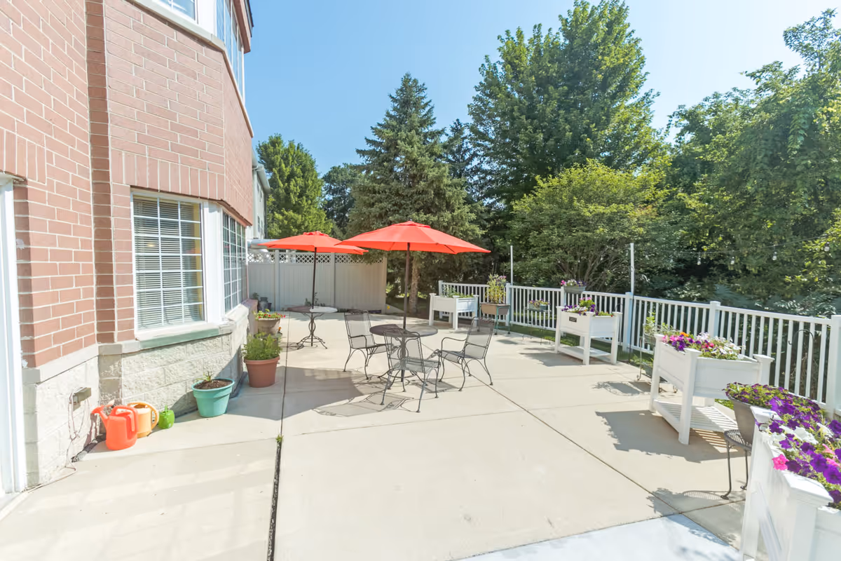 Sunlit outdoor patio with metal tables and red umbrellas, potted planters, and surrounding trees next to a brick building.