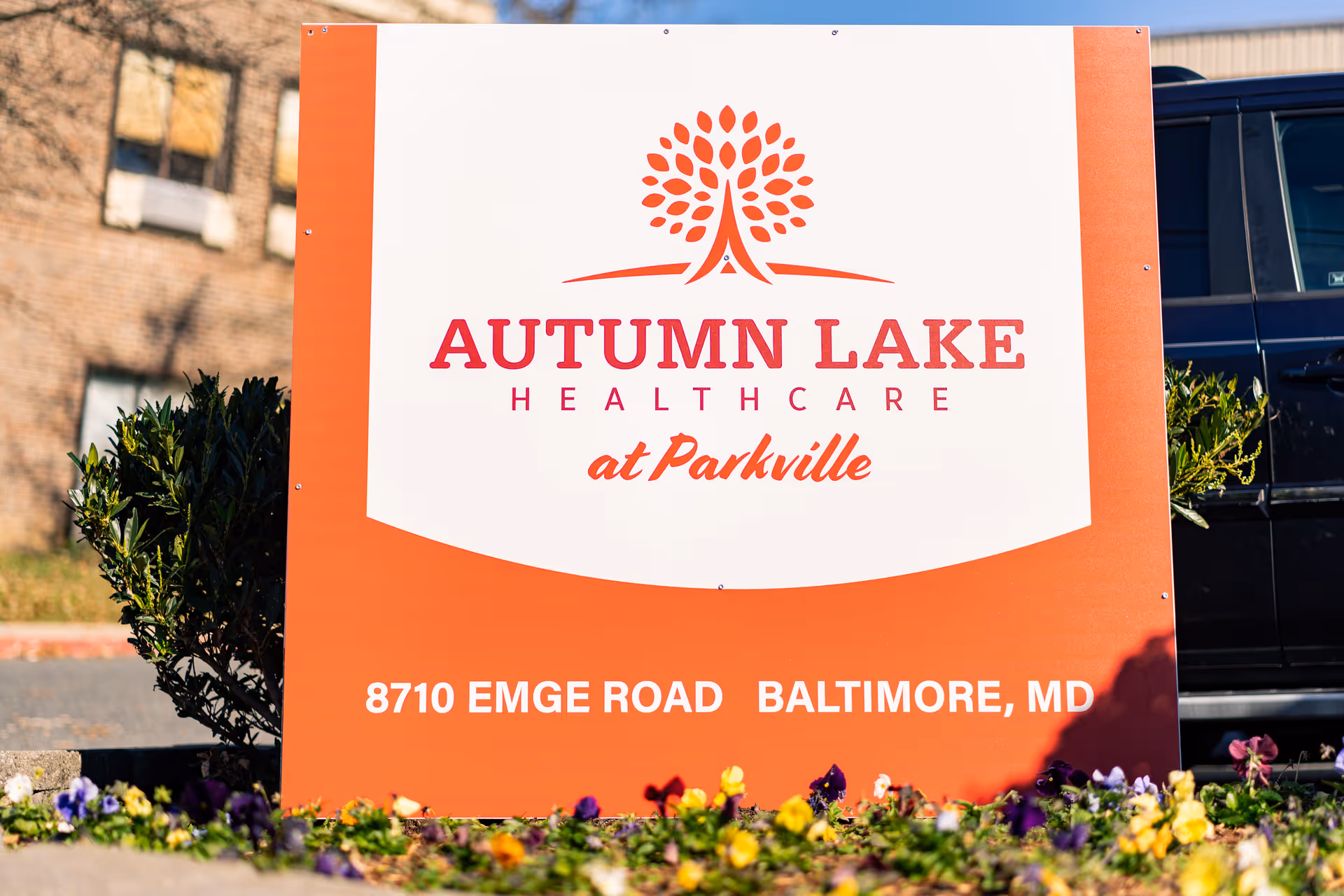 A large orange and white sign for Autumn Lake Healthcare at Parkville, located at 8710 Emge Road, Baltimore, MD, with flowers and greenery in the foreground and a building and vehicle in the background.