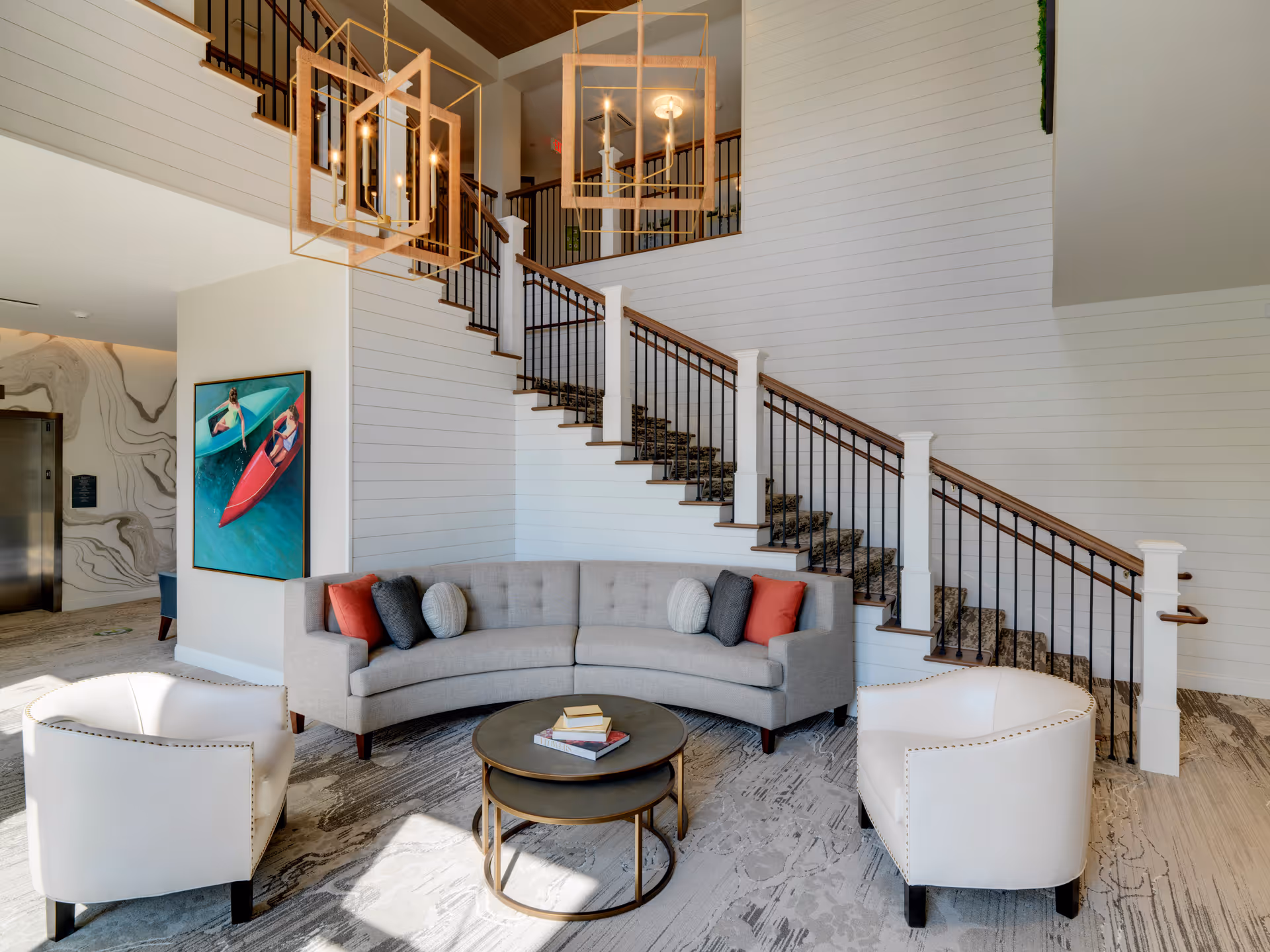 A modern and cozy seating area in a senior living facility featuring a curved gray sofa with multiple cushions, two white armchairs, and two round nested coffee tables with books on top. Behind the seating area is a staircase with a wooden handrail and black balusters, and two geometric pendant lights hang from the ceiling. A colorful painting of two people in kayaks is mounted on the wall near an elevator with a marble-patterned wall.