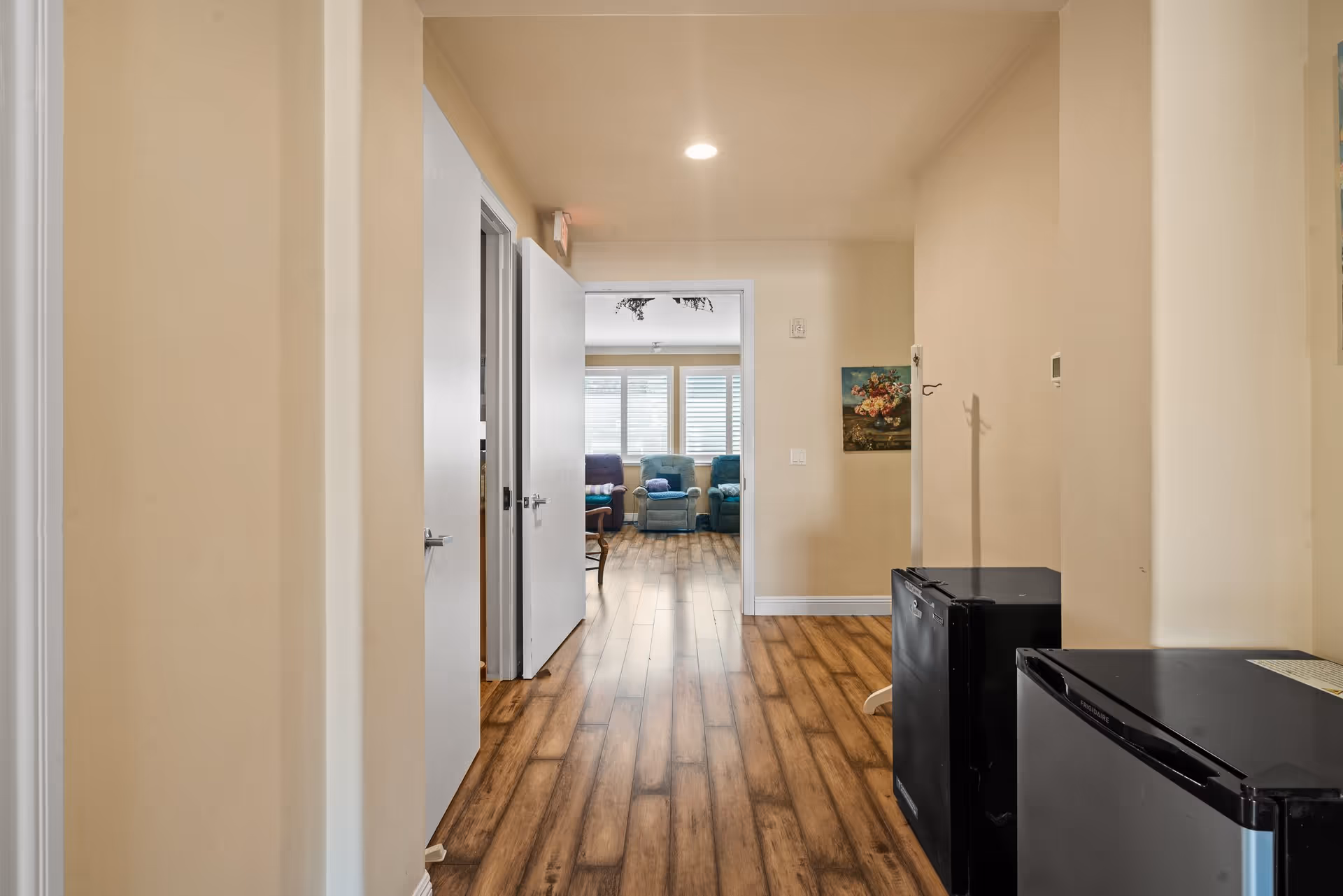 Hallway with wood-look flooring leading into a living area with recliners and shuttered windows.