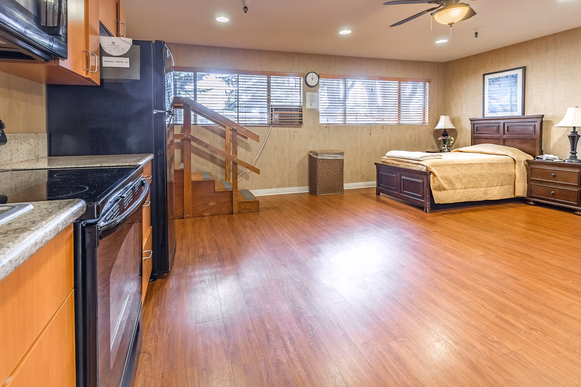 A spacious room with a wooden floor featuring a kitchen area on the left with a stove, oven, microwave, and refrigerator. On the right side, there is a bed with a wooden headboard and matching nightstands with lamps. A ceiling fan with a light is mounted above, and a set of windows with blinds is on the far wall above a small staircase and a wastebasket.