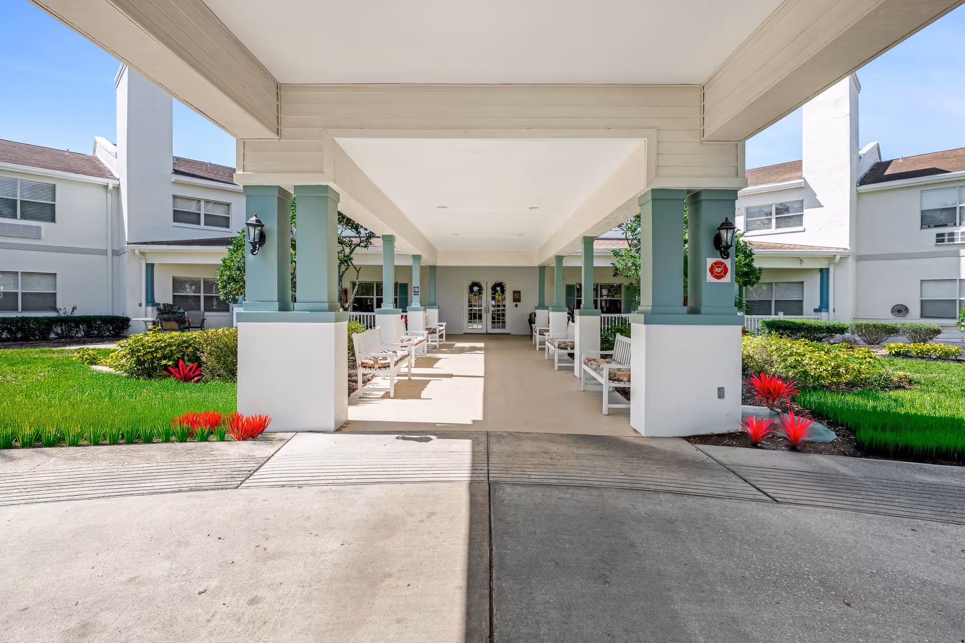 Covered entrance area of a senior living facility with white benches on both sides, green pillars, and a pathway leading to double glass doors. The building is white with multiple windows, surrounded by green grass and landscaped plants with red flowers.