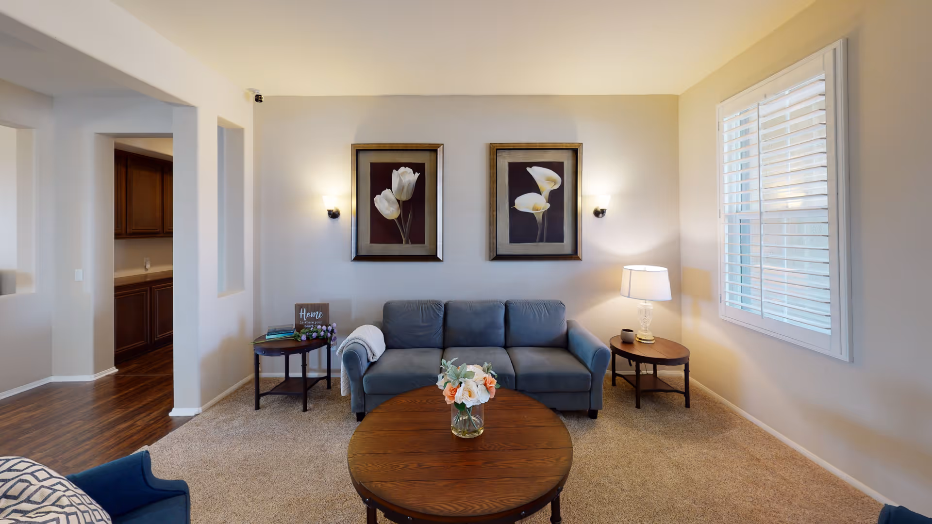 A cozy living room with a blue sofa centered against a beige wall, flanked by two framed floral paintings. There are two round wooden side tables on either side of the sofa, one with a lamp and the other with books and a decorative sign. A round wooden coffee table with a vase of flowers is in front of the sofa. The room has beige carpeting and a window with white shutters on the right side.