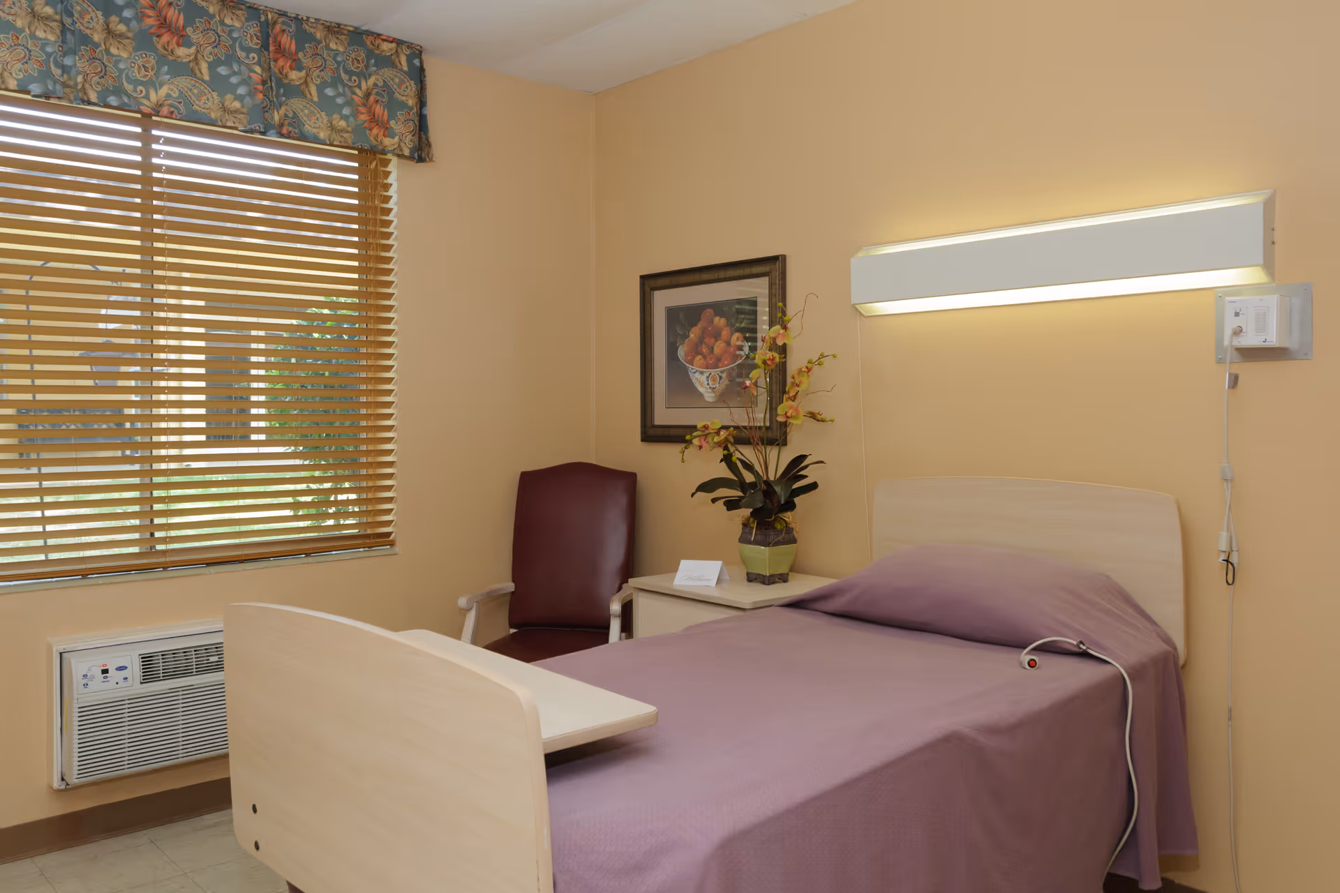A simple, clean bedroom in a senior living facility with a single bed covered in a purple blanket, a wooden headboard, a bedside table with a potted plant and a framed picture on the wall. There is a maroon chair next to the table, a window with wooden blinds and a floral valance, and an air conditioning unit below the window. A wall-mounted light fixture is above the bed.