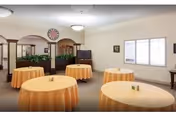 A dining room with five round tables covered in yellow tablecloths, each with a small centerpiece. The room has beige walls, a large window letting in natural light, a clock on the wall, and a wooden divider with plants behind it.