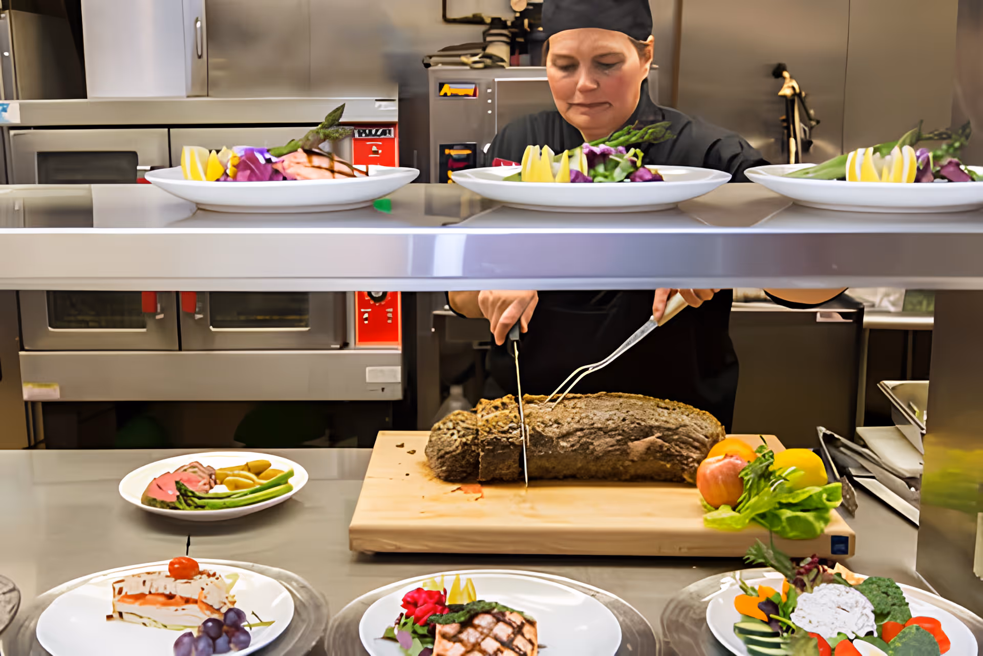 A chef in a commercial kitchen slicing a large piece of cooked meat on a wooden cutting board. Several plated dishes with vegetables, grilled salmon, and dessert are arranged on the counter in front of the chef. The kitchen has stainless steel appliances and equipment.