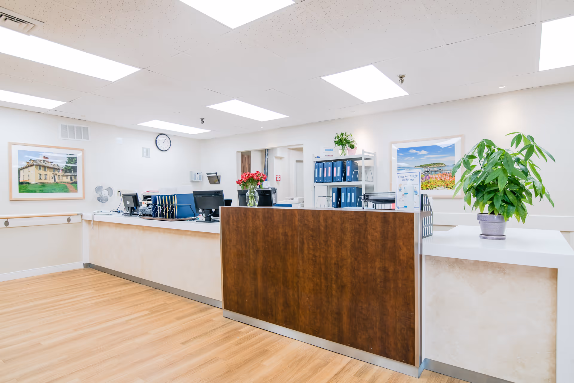 Reception desk area in a senior living facility with a wooden front panel and white countertop. The desk has computer monitors, office supplies, and a vase with red flowers. The room has light-colored walls, wood flooring, ceiling lights, framed pictures on the walls, a clock, and potted plants on the counter.