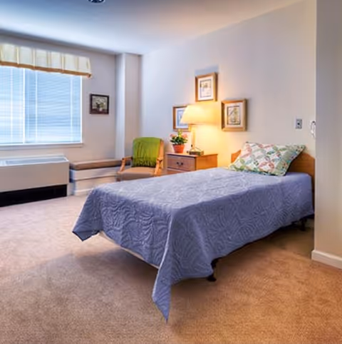 A cozy bedroom with a single bed covered in a blue quilt and a patterned pillow. Next to the bed is a wooden nightstand with a lamp and a small potted plant. There are three framed pictures hanging on the wall above the nightstand. A cushioned chair with a green throw is positioned near a window with blinds and a valance. The room has beige carpeting and light-colored walls.