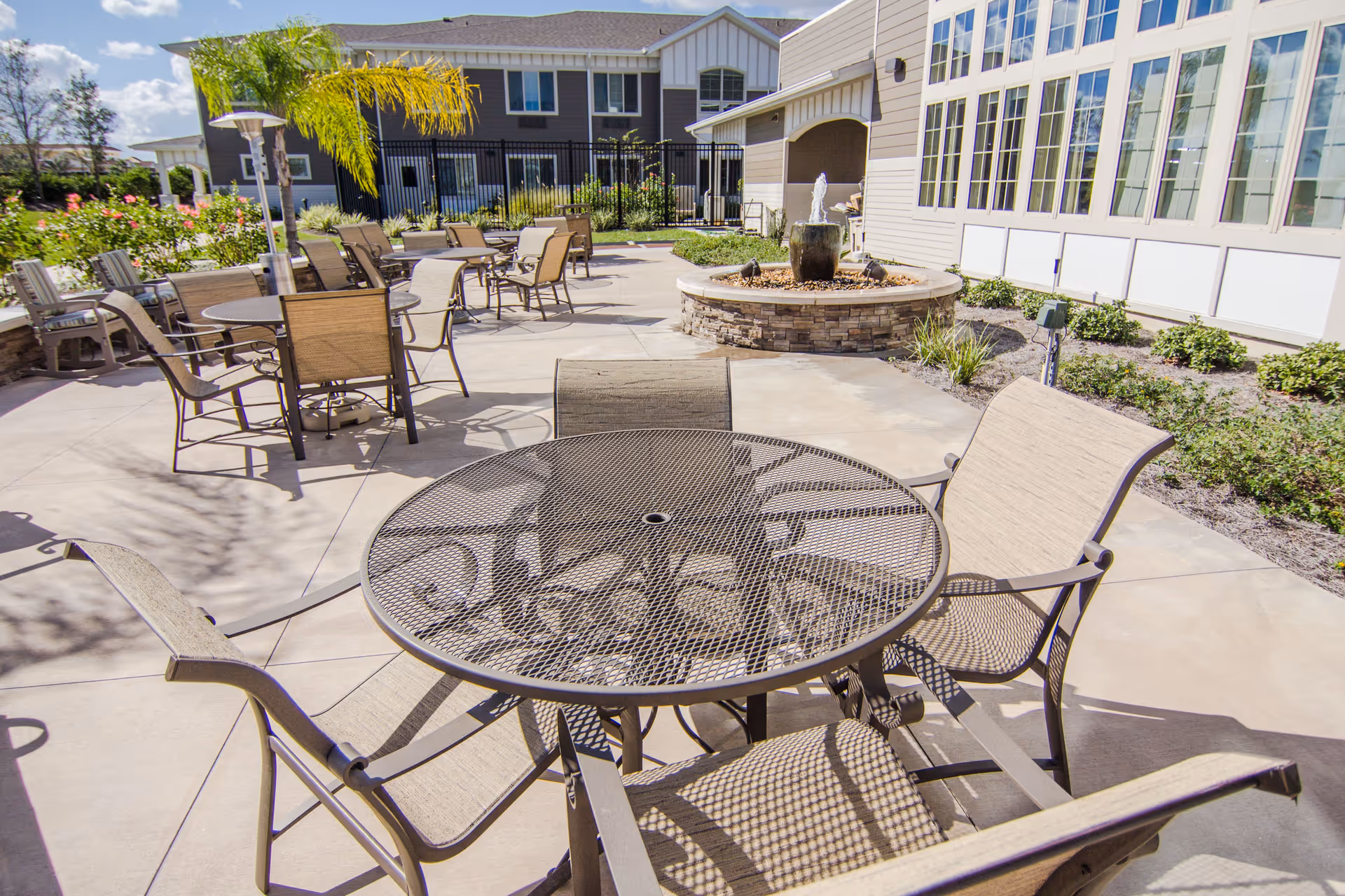 Outdoor patio area at The Willows featuring multiple metal mesh tables with beige cushioned chairs arranged around them. There is a circular stone water fountain in the background, surrounded by landscaping with green shrubs and palm trees. The building with large windows is visible on the right side under a clear blue sky.