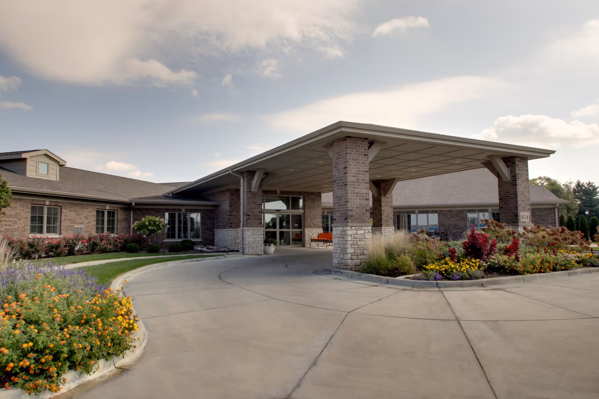 Exterior view of a senior living facility entrance with a covered drop-off area supported by brick pillars, surrounded by landscaped flower beds and a curved driveway under a partly cloudy sky.