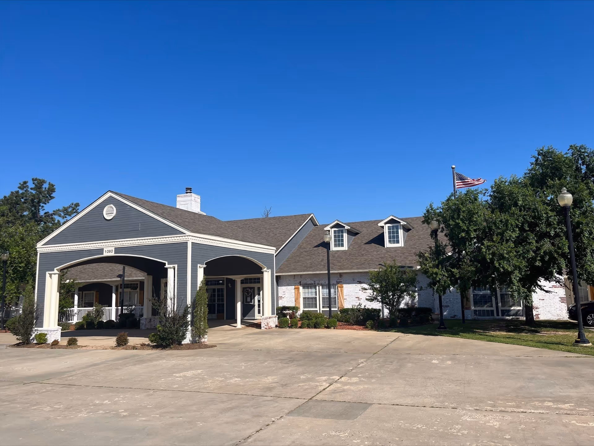 Front exterior of a single-story senior living facility with a covered entrance, American flag, and parking area under a clear blue sky.