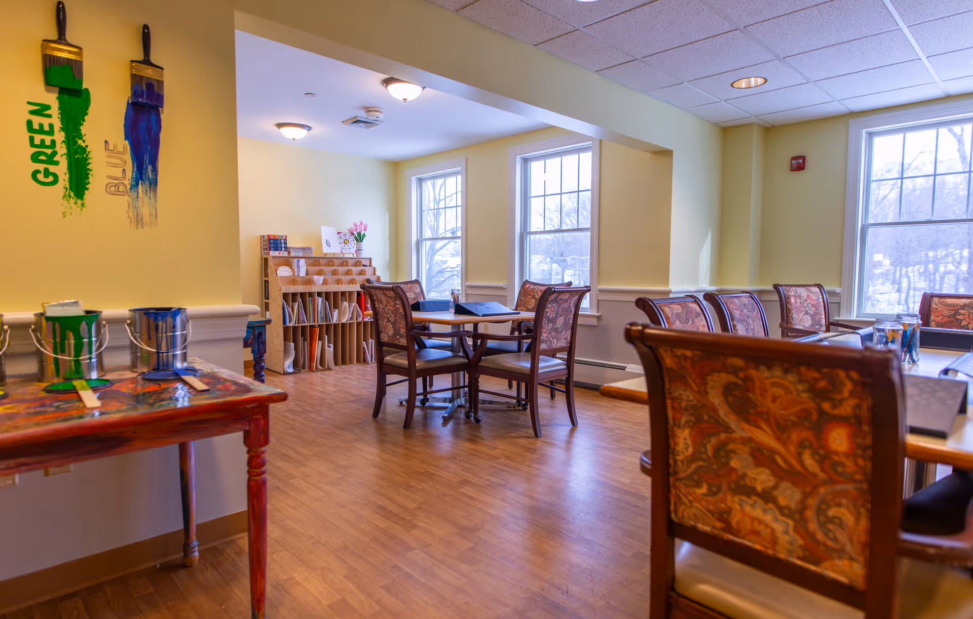 A bright room with wooden flooring and several tables surrounded by chairs with patterned upholstery. On the left side, there is a small table with paint cans and brushes, and a wall decoration featuring two paintbrushes with green and blue paint. The room has large windows letting in natural light and a wooden shelf with art supplies and books in the background.