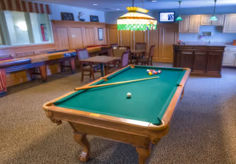 A recreational room featuring a pool table with billiard balls and cues, a shuffleboard table along the wall, several tables and chairs, wooden cabinetry, and a bar area with a countertop and pendant lights.