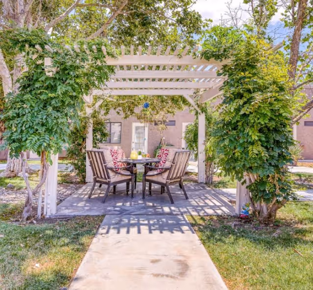 Outdoor seating area under a white pergola with green vines growing on the sides. Four cushioned chairs with patterned pillows surround a round table with a pitcher of lemonade and glasses. The area is paved and surrounded by grass and trees, with a building visible in the background.