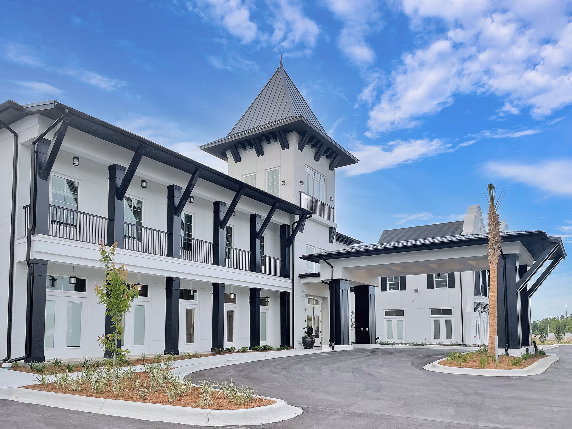 Exterior view of a modern senior living facility building with white walls and black accents, featuring a covered entrance, multiple windows, and a tower-like structure under a partly cloudy blue sky.
