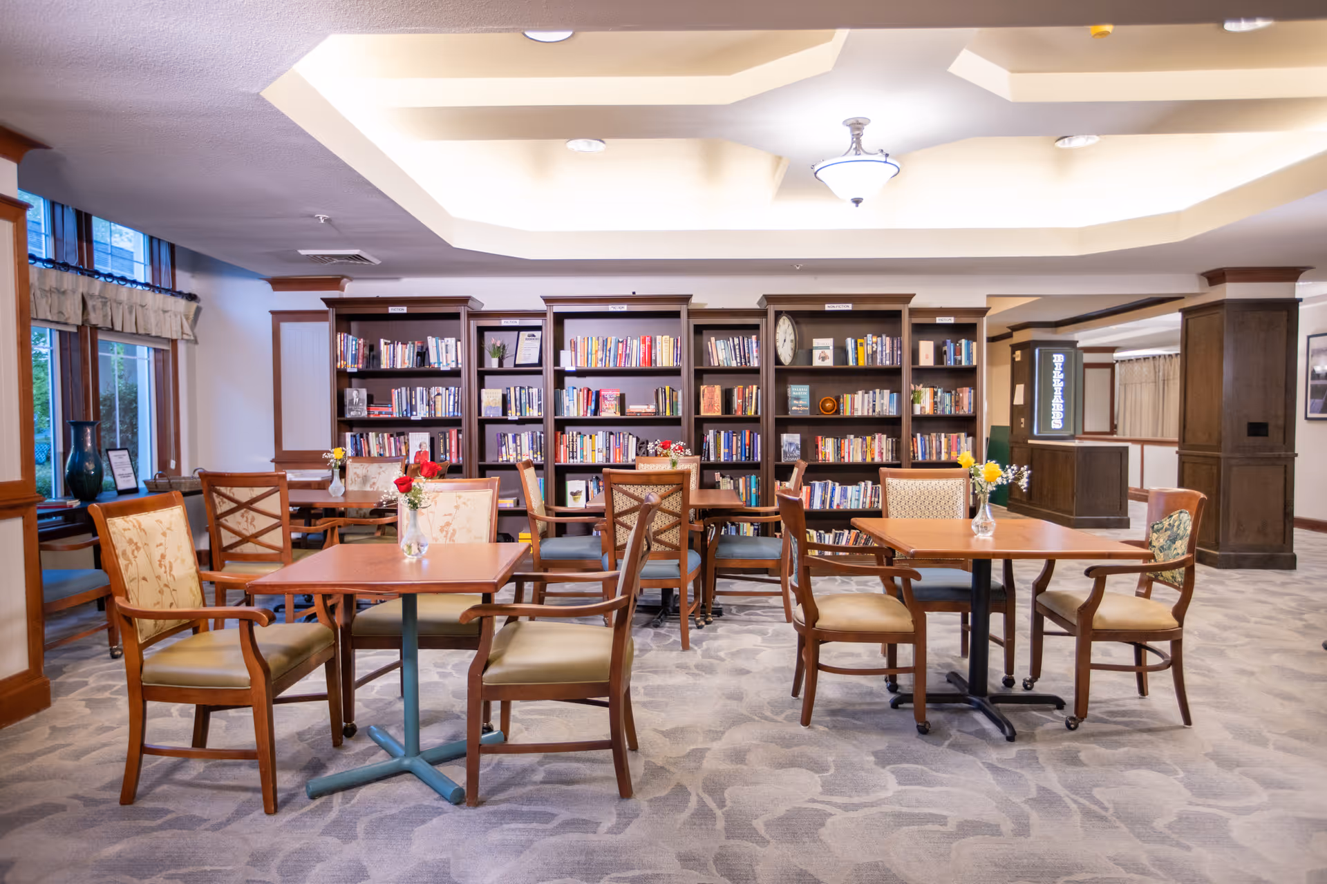 A well-lit common area with multiple wooden tables and cushioned chairs arranged on a patterned carpet. Behind the tables, there are tall bookshelves filled with books and decorative items. The ceiling features a recessed design with a central light fixture. Windows with valances are visible on the left side, and there are small vases with flowers on the tables.