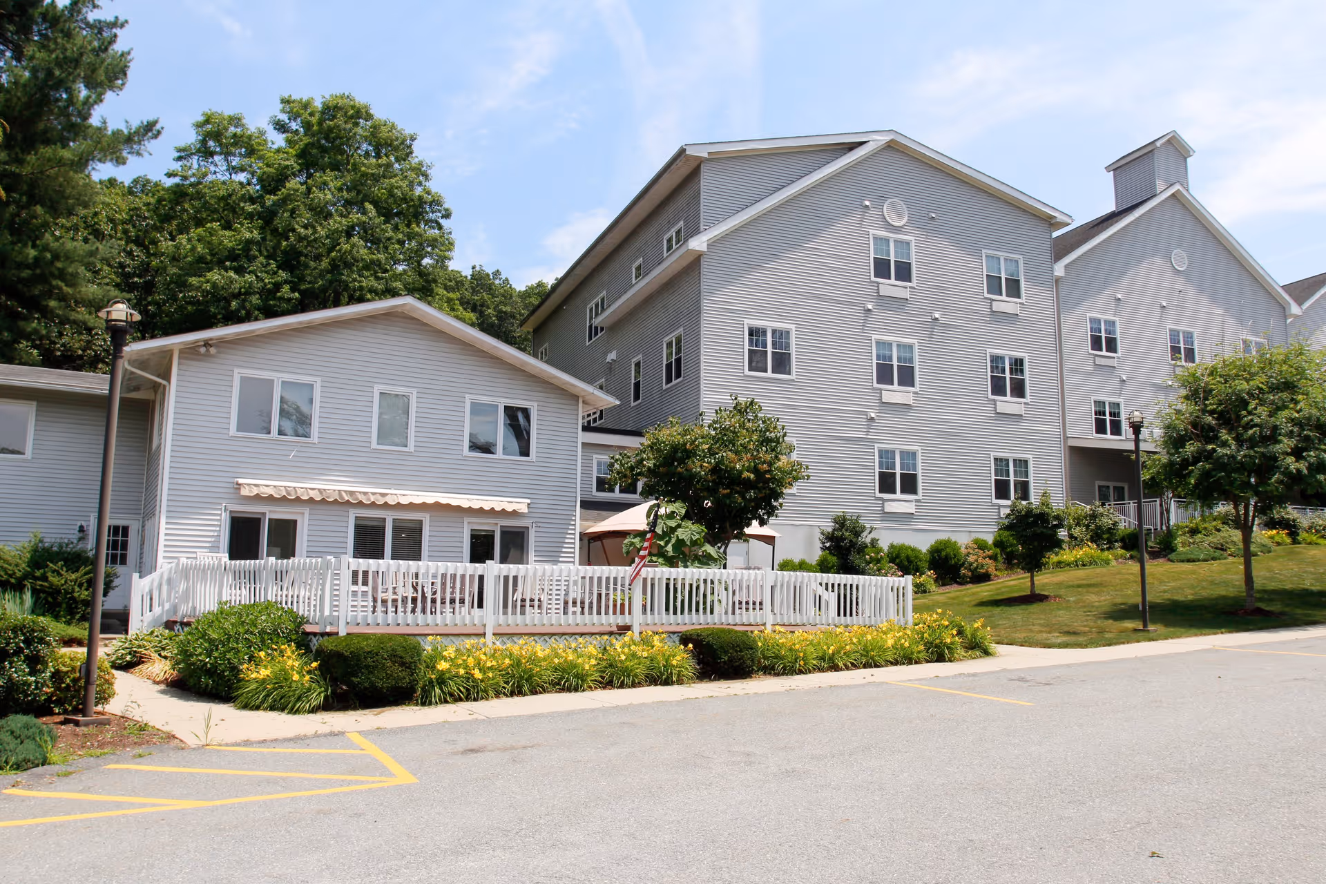 Exterior view of The Residence at Whitcomb House, showing a multi-story gray building with white trim, surrounded by green trees, shrubs, and a small garden with yellow flowers. There is a white fence enclosing a patio area and a parking lot in the foreground under a partly cloudy sky.
