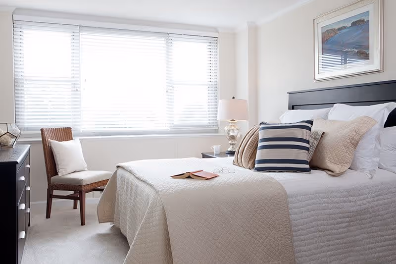 A bright bedroom with a large window covered by white blinds. The bed is neatly made with a beige quilt and multiple pillows in neutral and striped patterns. A wooden chair with a white cushion is placed near the window. A bedside table with a lamp and a cup is next to the bed, and a framed landscape painting hangs on the wall above the headboard.